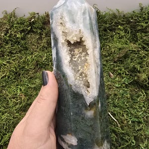 May include: A polished, green and white moss agate crystal tower with a geode cavity filled with sparkling crystals. The gemstone is held in a hand, showcasing its natural beauty and unique patterns. The tower is approximately 6 inches tall.