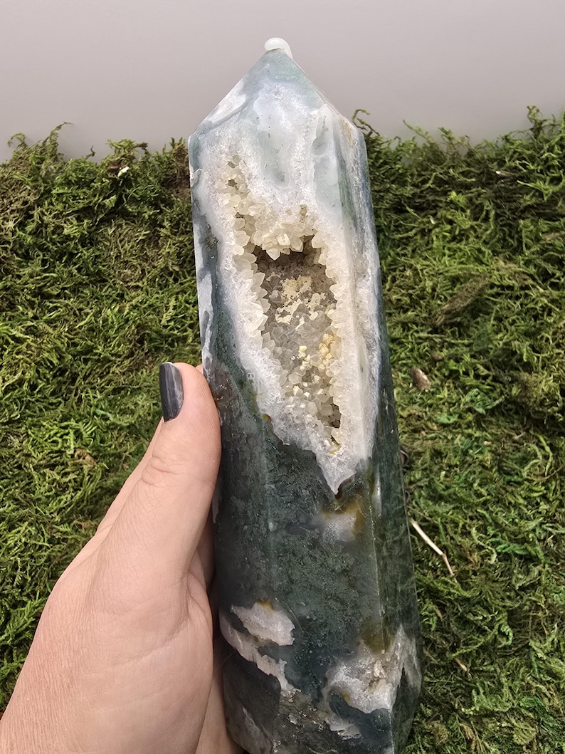 May include: A polished, green and white moss agate crystal tower with a geode cavity filled with sparkling crystals. The gemstone is held in a hand, showcasing its natural beauty and unique patterns. The tower is approximately 6 inches tall.