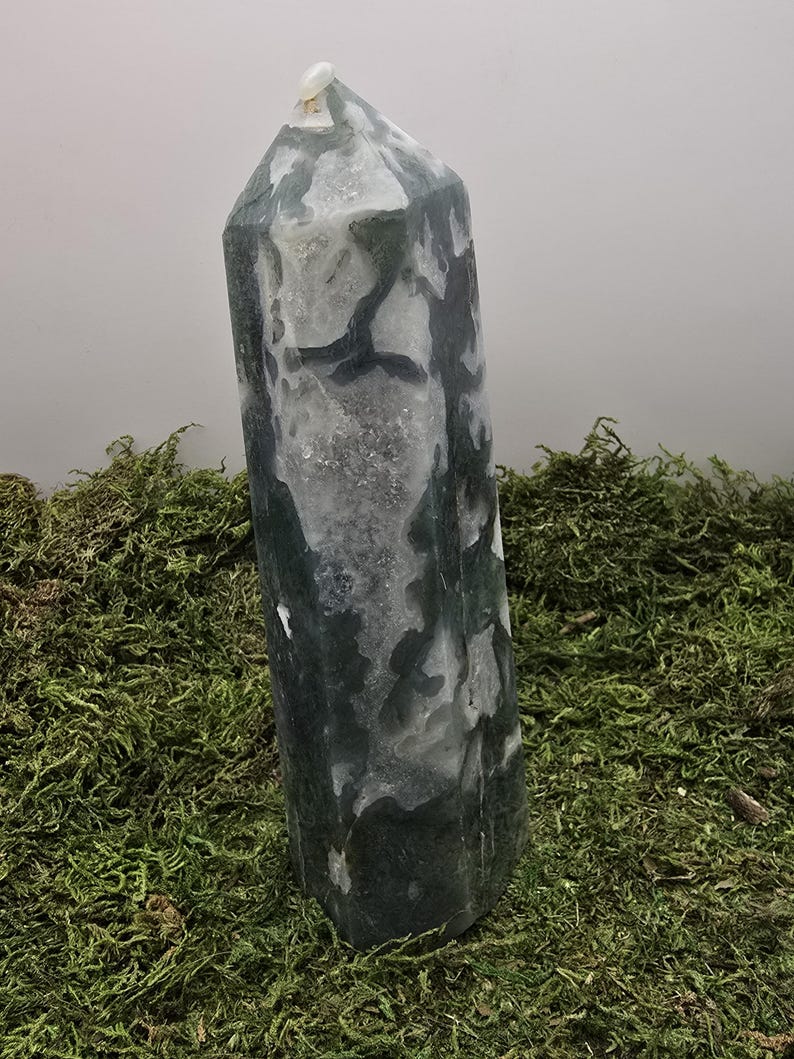 May include: A tall, polished crystal obelisk with a mottled pattern of dark green and white. The crystal is standing upright on a bed of green moss, with a light gray background.