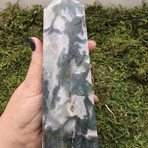 May include: A tall, rectangular crystal obelisk with a polished surface. The stone features a mix of white, gray, and green patterns. The obelisk is held against a backdrop of green moss.