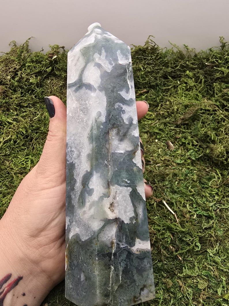 May include: A tall, rectangular crystal obelisk with a polished surface. The stone features a mix of white, gray, and green patterns. The obelisk is held against a backdrop of green moss.