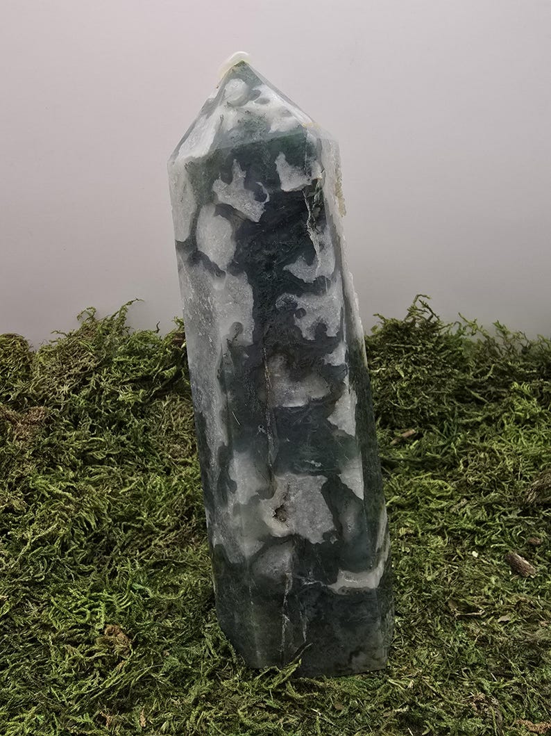 May include: A tall, polished crystal obelisk with a pointed top. The stone features a pattern of dark green and white inclusions. The obelisk is displayed on a bed of green moss, creating a natural aesthetic.