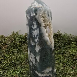 May include: A tall, polished crystal obelisk with a pointed top. The stone features a mix of dark green and white patterns, with a cavity of small, light-colored crystals. The obelisk is displayed on a bed of green moss.
