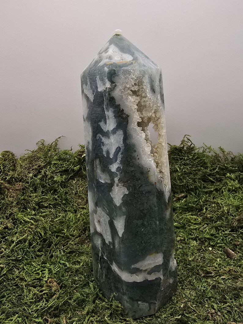 May include: A tall, polished crystal obelisk with a pointed top. The stone features a mix of dark green and white patterns, with a cavity of small, light-colored crystals. The obelisk is displayed on a bed of green moss.