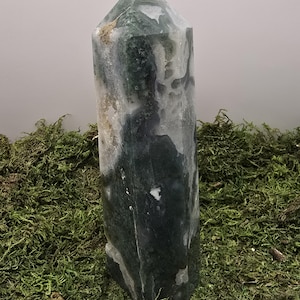 May include: A tall, polished crystal tower with a pointed top. The stone features a mix of green, white, and gray colors, with a small, light-colored stone at the apex. The crystal is displayed on a bed of green moss.