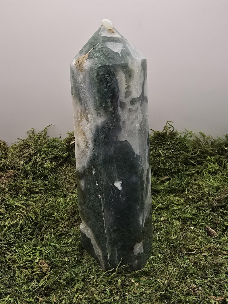 May include: A tall, polished crystal tower with a pointed top. The stone features a mix of green, white, and gray colors, with a small, light-colored stone at the apex. The crystal is displayed on a bed of green moss.