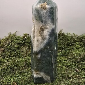 May include: A tall, polished crystal obelisk with a faceted top. The crystal features a mix of translucent white, dark green, and gray patterns. A section of the crystal has a rough, white, and beige interior. The obelisk is displayed on a bed of green moss.