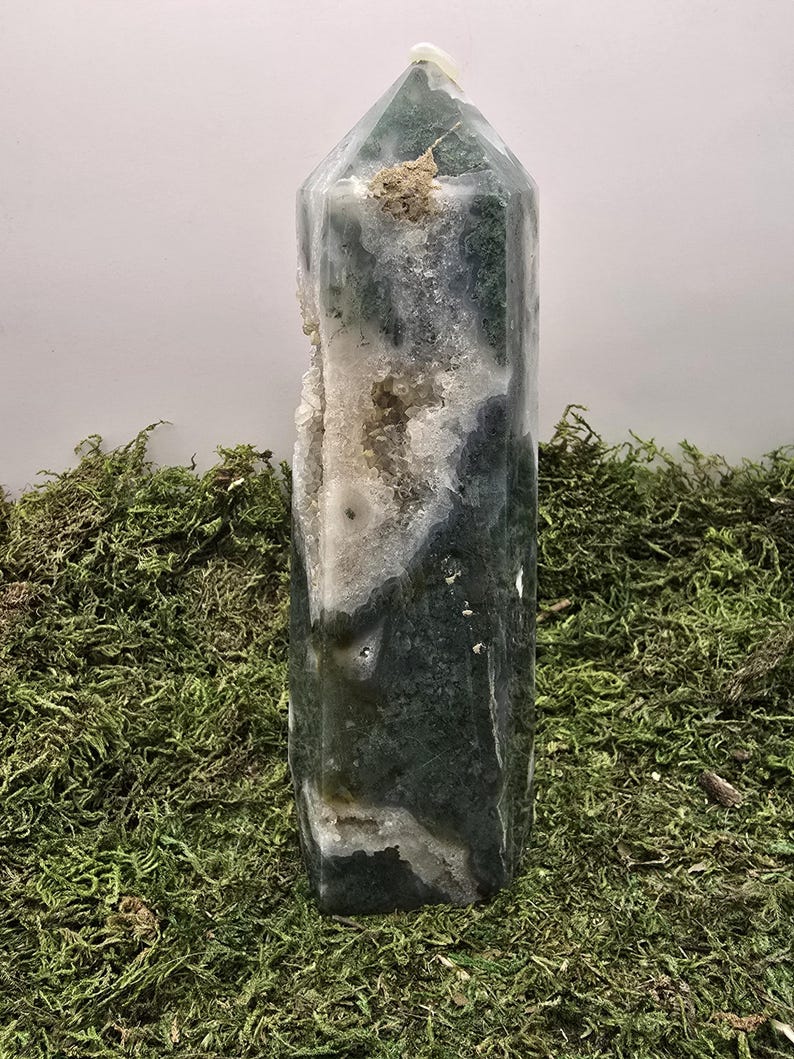 May include: A tall, polished crystal obelisk with a faceted top. The crystal features a mix of translucent white, dark green, and gray patterns. A section of the crystal has a rough, white, and beige interior. The obelisk is displayed on a bed of green moss.