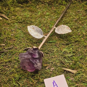 May include: A handcrafted, purple fluorite rose with a silver stem and leaves. The rose is set against a backdrop of green moss. A small white card with the letter "A" is in the lower right corner.