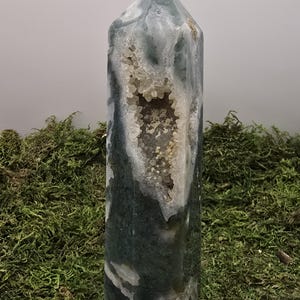 May include: A tall, polished crystal obelisk with a geode cavity. The crystal features a mix of green, white, and gray hues, with a cluster of small, sparkling crystals inside the geode. The obelisk is set against a backdrop of green moss.
