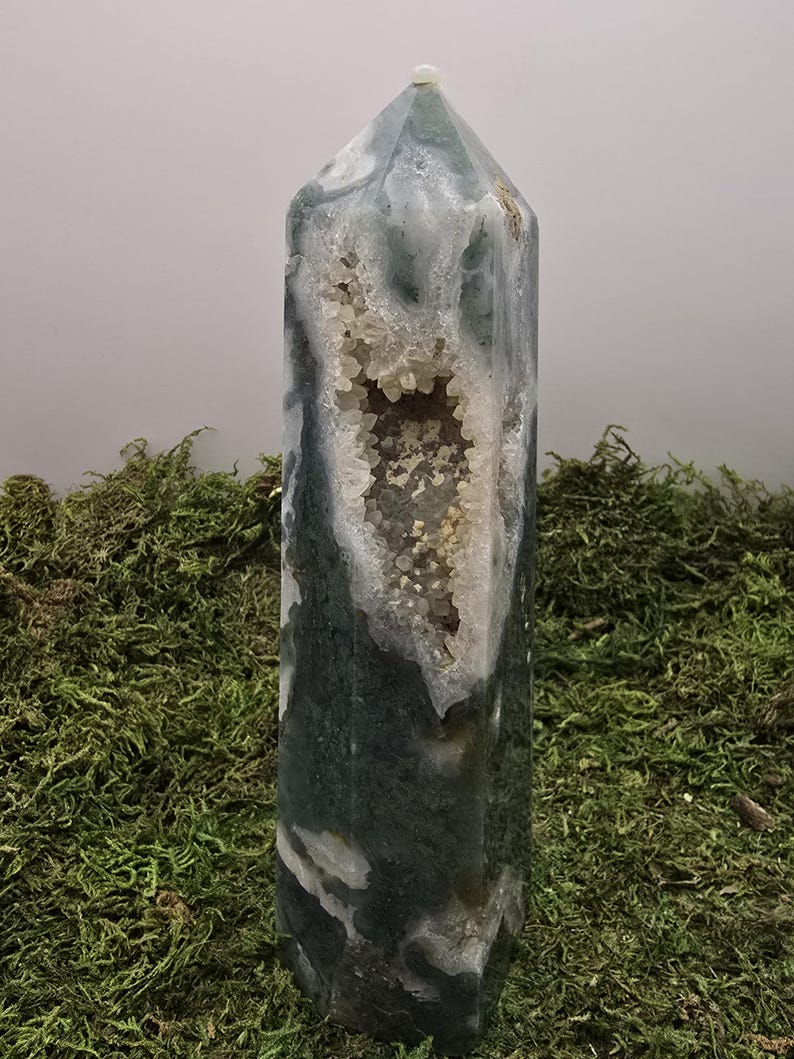 May include: A tall, polished crystal obelisk with a geode cavity. The crystal features a mix of green, white, and gray hues, with a cluster of small, sparkling crystals inside the geode. The obelisk is set against a backdrop of green moss.