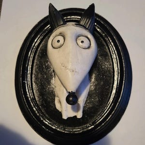 May include: A white and black dog-like sculpture with large, round eyes and pointed ears. The sculpture has a black nose and is set against a black oval frame. The artwork has a whimsical, cartoonish style.