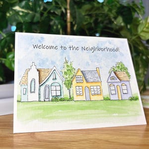 A blank greeting card featuring a watercolor and ink print of three whimsical houses painted in pastel colors sitting in a row. A green grass fills the foreground while two trees and a patchy blue sky fill the background. The words, "Welcome to the neighborhood!" are written across the sky in a playful font. Card measures 5.5 inches long by 4.25 inches tall.
