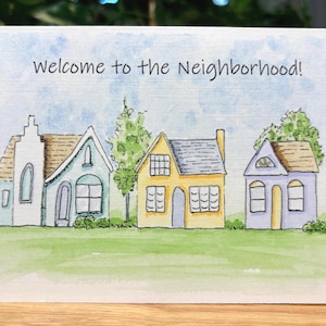 May include: A greeting card featuring the text "Welcome to the Neighbourhood!" above a watercolour illustration of houses. The houses are in various colours, set on a green lawn under a blue sky.