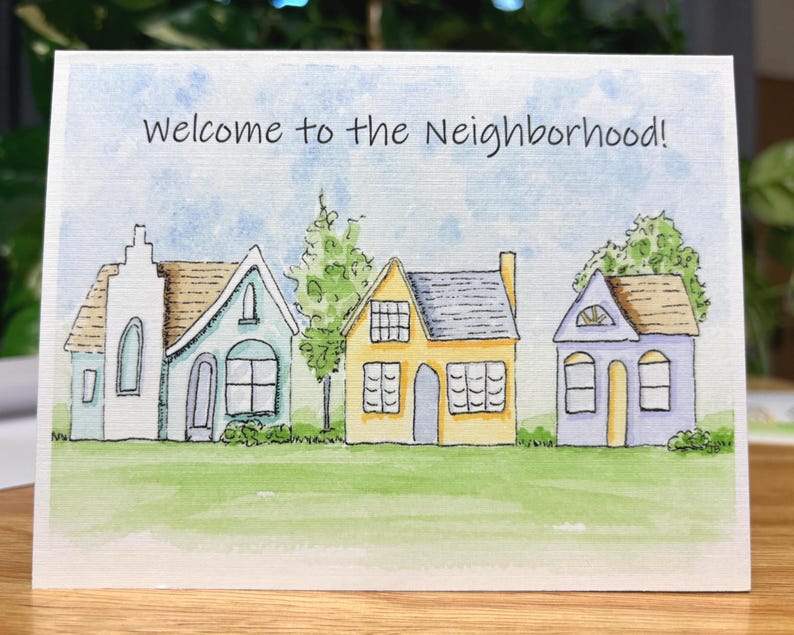 May include: A greeting card featuring the text "Welcome to the Neighbourhood!" above a watercolour illustration of houses. The houses are in various colours, set on a green lawn under a blue sky.