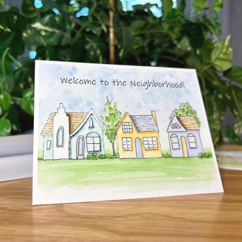 A blank greeting card featuring a watercolor and ink print of three whimsical houses painted in pastel colors sitting in a row. A green grass fills the foreground while two trees and a patchy blue sky fill the background. The words, "Welcome to the neighborhood!" are written across the sky in a playful font. Card measures 5.5 inches long by 4.25 inches tall.
