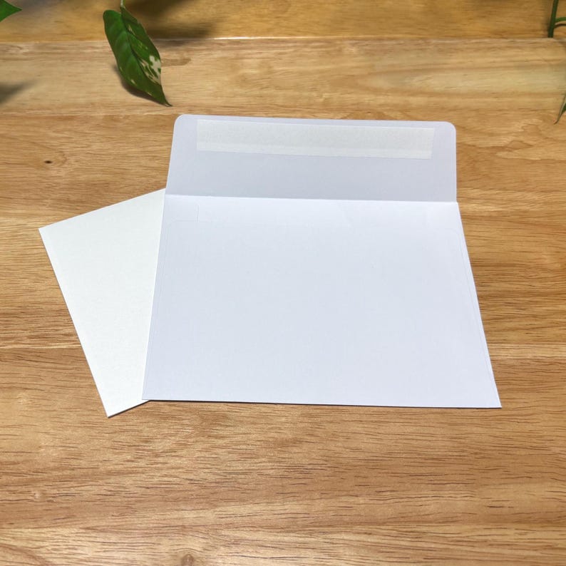 May include: Two white envelopes, one open, are displayed on a wooden surface. The open envelope reveals its interior, and the other lies beside it. A leaf is visible in the background.
