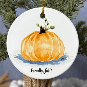May include: Round white ceramic ornament featuring a watercolor painting of an orange pumpkin with green vines. The text "Finally, fall!" is printed below the pumpkin. A gold string is attached for hanging.