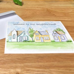 May include: A watercolour greeting card with the text "Welcome to the Neighbourhood!" featuring three colourful houses on a green lawn. A white envelope is visible in the background.