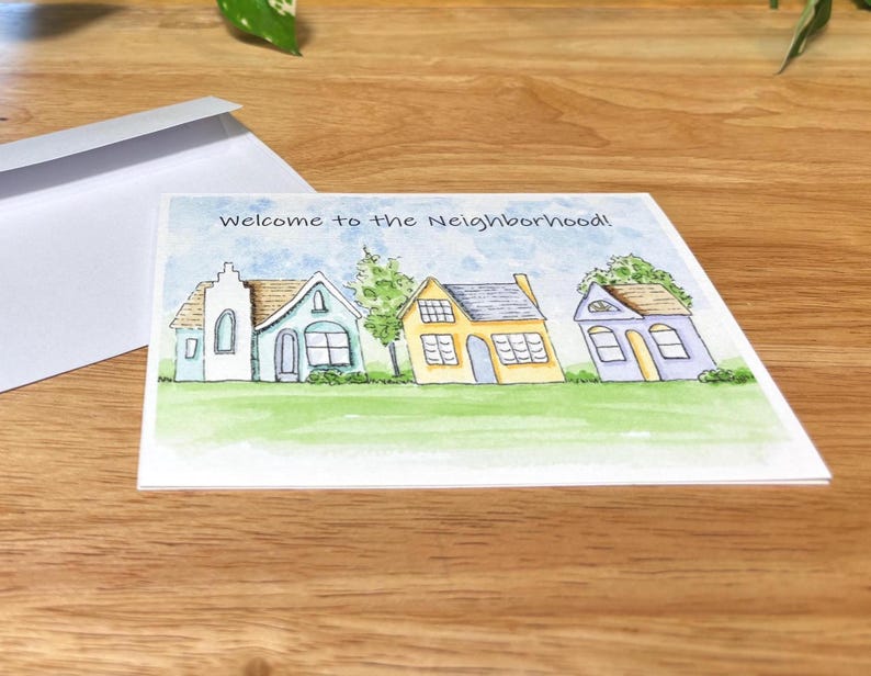 May include: A watercolour greeting card with the text "Welcome to the Neighbourhood!" featuring three colourful houses on a green lawn. A white envelope is visible in the background.