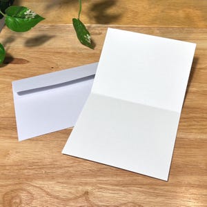 May include: A blank white greeting card is open, showing a clean interior. A matching white envelope sits nearby on a wooden surface. Green plant leaves are visible in the background, adding a natural touch to the scene.