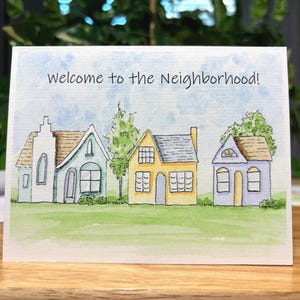 May include: A greeting card featuring a watercolour illustration of four houses on a green lawn beneath a blue sky. The text "Welcome to the Neighbourhood!" is written across the top.