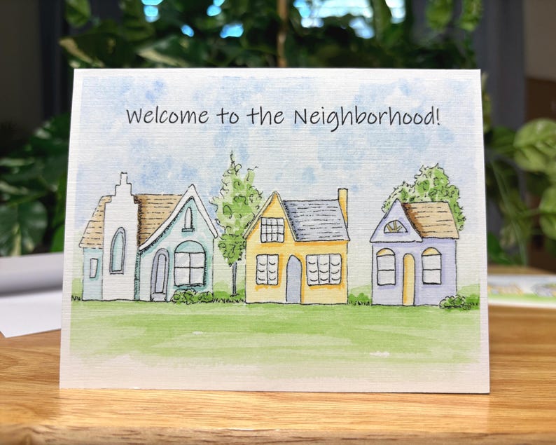 May include: A greeting card featuring a watercolour illustration of four houses on a green lawn beneath a blue sky. The text "Welcome to the Neighbourhood!" is written across the top.