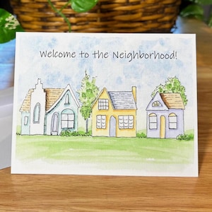 May include: A watercolour greeting card with the text "Welcome to the Neighbourhood!" shows three houses. The houses are white, blue, and yellow, with green grass and trees. The sky is light blue.