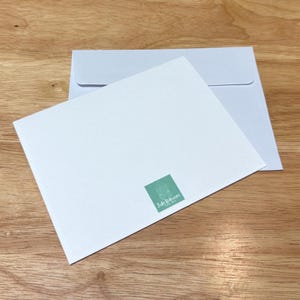 May include: A white greeting card and envelope set on a wooden surface. The card has a small, square, green logo in the bottom right corner. The envelope is light blue.