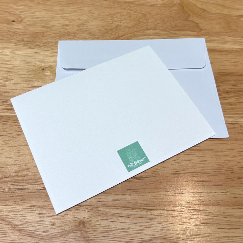 May include: A white greeting card and envelope set on a wooden surface. The card has a small, square, green logo in the bottom right corner. The envelope is light blue.