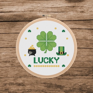 May include: A cross-stitch embroidery hoop featuring a green four-leaf clover, a pot of gold, and a green hat. The word "LUCKY" is stitched in green below the symbols, with additional green shamrocks and yellow dots.
