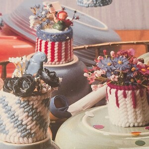 May include: Three small cylindrical containers crafted from plastic canvas with yarn stitching. One features red and white stripes with a small flag, another has a blue and white diagonal pattern with a bird, and the third has white yarn with red drips.