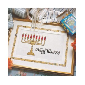 May include: Cross-stitch artwork of a gold menorah with red candles, framed in gold. The words "Happy Hanukkah" are stitched in black script below the menorah. A blue box with a menorah design is visible.