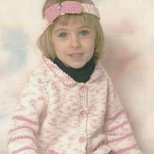 May include: A knitted cardigan featuring a white and pink variegated yarn pattern with a button-down front and a matching knitted headband with a central bow detail. The garment has long sleeves and a rounded collar, showcasing a soft texture.