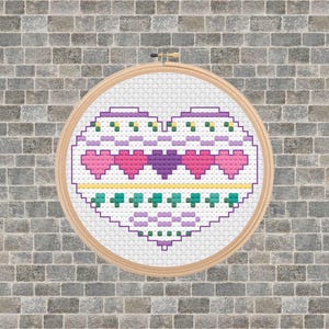 May include: A cross-stitch heart design in a wooden embroidery hoop. The heart features pink, purple, and green cross-stitch patterns. The background is white, and the hoop is set against a gray brick wall.
