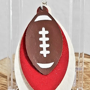 May include: A pair of layered leather earrings with a white football design on the top layer. The bottom layer is white, the middle layer is red, and the top layer is brown.