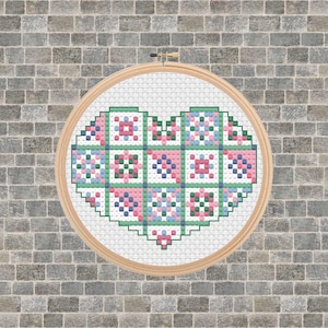 May include: A cross-stitch heart design in a wooden embroidery hoop. The heart is composed of pink, blue, and green squares, set against a white background. The hoop is displayed against a gray brick wall.