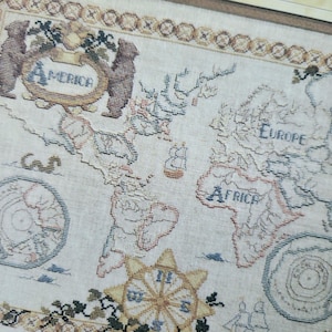 May include: A detailed, vintage-style map featuring the continents of the world. The map is embroidered on a cream-colored fabric, with the continents outlined in brown and labeled with text. Decorative elements include bears, compasses, and a border.