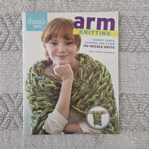 May include: A book cover for "Arm Knitting" by Linda Zemba Burhance. The cover features a woman wearing a green and yellow arm-knitted cowl. The book title is in large, colorful letters, with the subtitle "Chunky Cowls, Scarves, and Other No-Needle Knits."