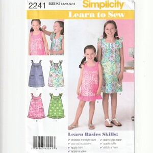 May include: Simplicity sewing pattern 2241 for children's dresses in sizes K5 7, 8, 10, 12, 14. The envelope features illustrations and photographs of four dress variations with different patterns, including floral and geometric prints, with ruffle details.