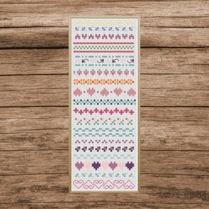 May include: A framed cross-stitch sampler with various colorful patterns. The design includes rows of geometric shapes, hearts, and floral motifs in shades of pink, blue, and green. The sampler is set against a light background.