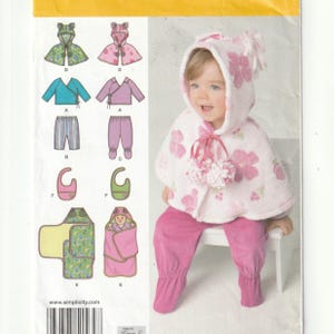 May include: A sewing pattern envelope featuring illustrations of baby clothing including capes, wrap tops, pants, bibs, and swaddles. A photograph shows a child wearing a white hooded cape with pink floral patterns and pink footed pants.