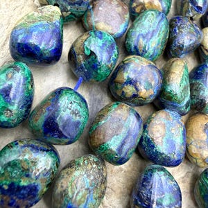 May include: A collection of polished, tumbled azurite-malachite beads featuring a vibrant mix of deep royal blue, forest green, and earthy tan mineral inclusions. Each bead has a smooth, rounded shape and is strung together on a thin blue cord.