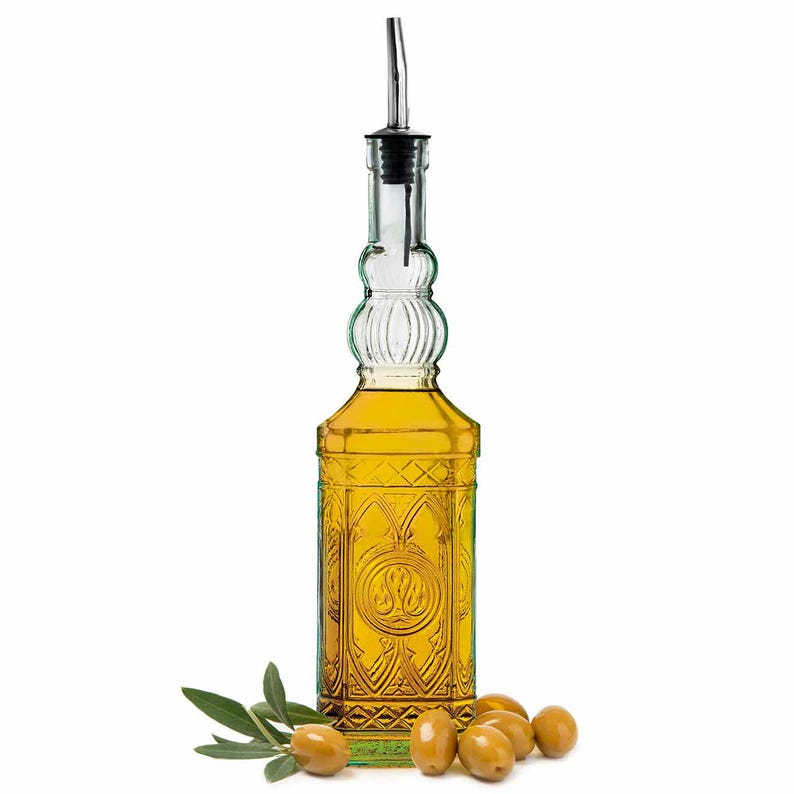 Op de afbeelding: Een heldere glazen olijfoliefles met een roestvrijstalen tuit, gevuld met gele olie. De fles heeft een sierlijk ontwerp en wordt vergezeld door olijven en een takje bladeren, wat wijst op een culinair thema.