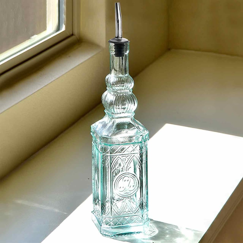 Op de afbeelding: Een heldere glazen fles met een decoratief ontwerp, voorzien van een zilveren tuit en een zwarte stopper. De fles heeft een sierlijke, vintage-ge&iuml;nspireerde vorm met een rechthoekige basis en een gedetailleerd patroon. De fles is ongeveer 25 cm hoog.