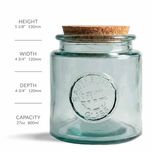 May include: A clear, turquoise-colored glass jar with a cork lid. The jar is cylindrical with a rounded base and a circular embossed design that reads "AUTHENTIC 100% RECYCLED GLASS". The jar is 5 1/8" high, 4 3/4" wide, and 4 3/4" deep, with a capacity of 27oz.
