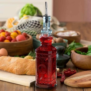 Op de afbeelding: Een decoratieve glazen fles gevuld met een rode vloeistof, met een sierlijk ontwerp. De fles heeft een zilveren schenktuit en is omgeven door verse producten, waaronder tomaten, brood en kruiden. De fles is ongeveer 25 cm hoog.