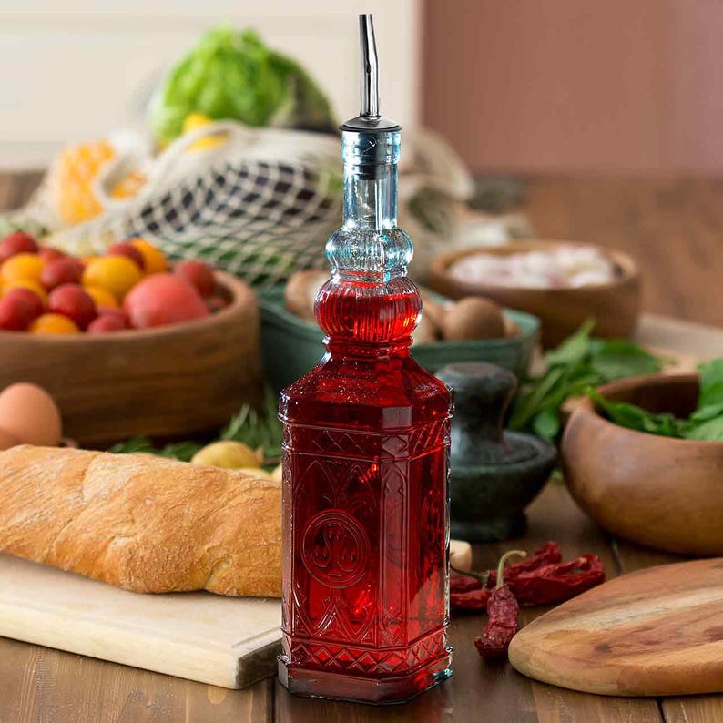 Op de afbeelding: Een decoratieve glazen fles gevuld met een rode vloeistof, met een sierlijk ontwerp. De fles heeft een zilveren schenktuit en is omgeven door verse producten, waaronder tomaten, brood en kruiden. De fles is ongeveer 25 cm hoog.