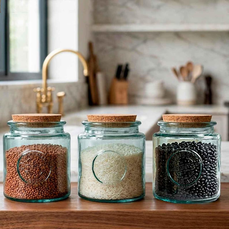 May include: Three glass jars with cork lids, each filled with different grains. The jars have a light blue tint and a circular design on the front. The jars are filled with brown lentils, white rice, and black beans.