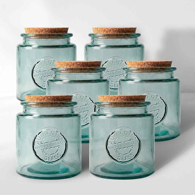 May include: Six clear, square glass jars with cork lids. The jars have a light blue tint and a circular embossed design that reads "AUTHENTIC 100% RECYCLED GLASS". The jars are arranged in a group.